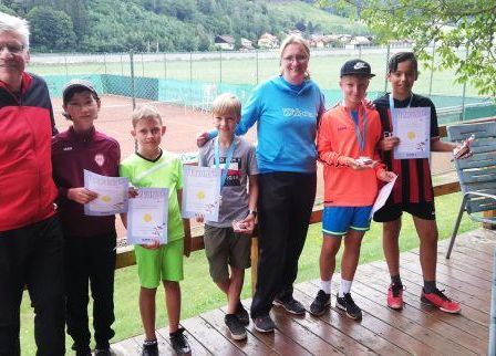 Sechs Kinder und zwei Erwachsene stehen auf einer Holzveranda und halten Urkunden in den Händen, ein Tennisplatz ist im Hintergrund zu sehen.