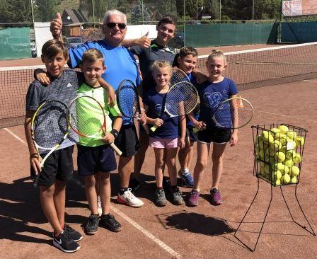 Eine Gruppe von Kindern und ein älterer Mann posieren für ein Foto auf einem Tennisplatz mit Schlägern und Tennisbällen.
