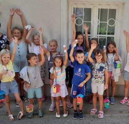 Eine Gruppe von Kindern, begleitet von einer Erwachsenen, posiert für ein Foto vor einem Gebäude mit einer Glastür. Sie halten kleine Vogelhäuschen.