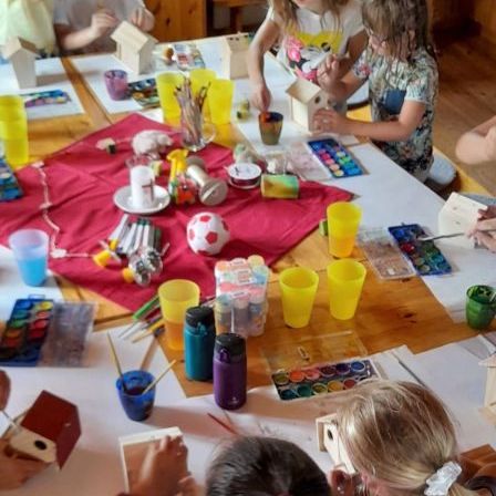 Eine Gruppe junger Mädchen sitzt um einen holzernen Tisch herum und ist mit einer Bastelaktivität beschäftigt. Jedes Kind hat ein kleines hölzerne Vogelhäuschen und malt es. Der Tisch ist mit verschiedenen Bastelutensilien wie Pinsel, Becher und Malpaletten gefüllt.