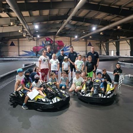 Eine Gruppe von Kindern und Erwachsenen posiert für ein Foto auf einer Go-Kart-Bahn. Zwei Go-Karts stehen auf der Strecke.