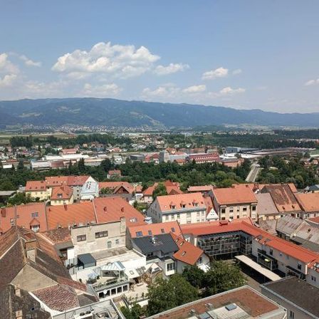 Luftaufnahme einer Stadt mit vielen Gebäuden, roten Dächern und Bäumen. Berge und ein klarer Himmel in der Ferne.