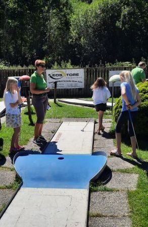 Eine Gruppe junger Frauen spielt Minigolf im Freien, wobei eine sich darauf vorbereitet, den Ball zu schlagen. Der Kurs hat ein blaues Loch mit dem Wort 'Verboten' darauf.