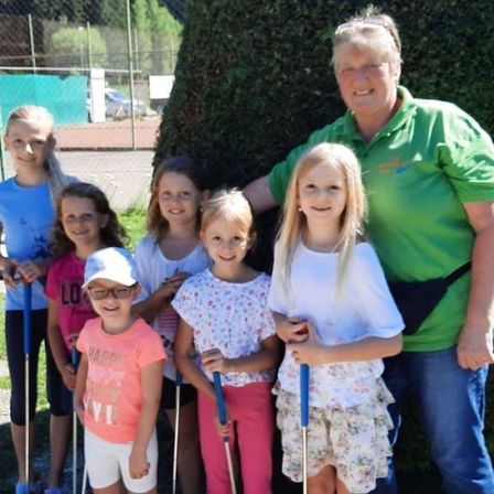 Eine Gruppe junger Mädchen und eine Frau mit grünem Polo posieren für ein Foto. Sie halten Golfschläger und hinter ihnen steht ein großer Busch.