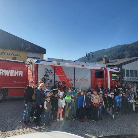 Eine Gruppe von Menschen, darunter Kinder, posiert für ein Foto vor einem roten Feuerwehrauto. Ein Gebäude mit dem Schriftzug 'und Festsaal' steht dahinter.