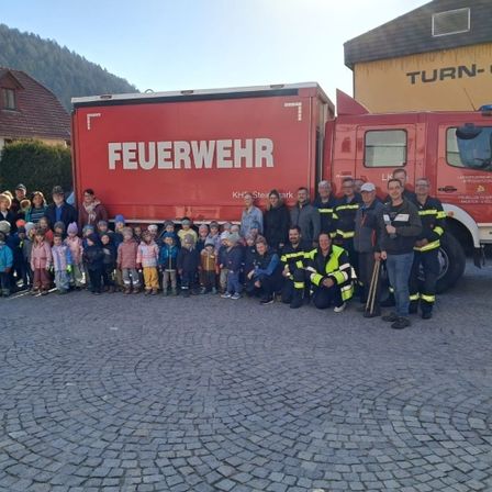 Eine Gruppe von Feuerwehrleuten und Kindern steht vor einem roten Feuerwehrauto in einem Dorf. Hinter ihnen befinden sich Gebäude und eine Bergkette.