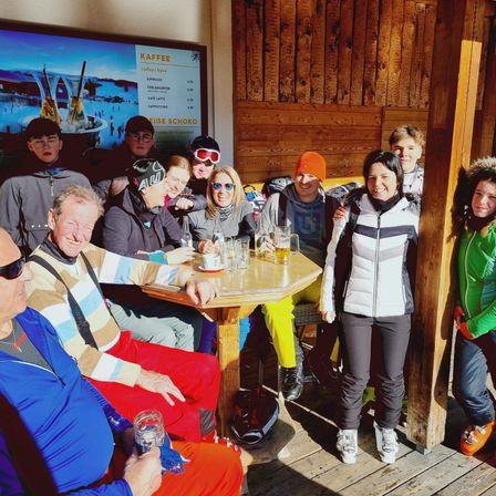 Eine Gruppe von Menschen versammelt sich drinnen und lächelt, einige mit Getränken. Eine Holzwand hat ein Schild für Kaffee. Ein Monitor zeigt ein Bild von Menschen auf einer Eisbahn.