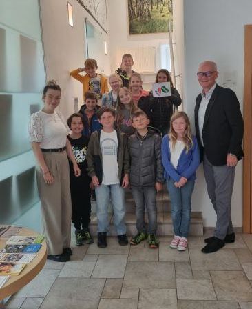 Eine Gruppe von Kindern und zwei Erwachsenen stehen zusammen auf einem gefliesten Boden. Der Mann hält ein Schild und die Frau ein Stück Papier. Dahinter befindet sich eine Treppe mit einem Gemälde.