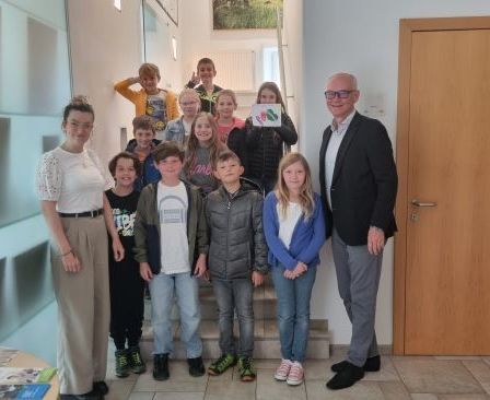 Eine Gruppe von Kindern und Erwachsenen steht auf einer Treppe in einem Raum. Ein Mann in formeller Kleidung und eine Frau in einem weißen Shirt stehen vorne. Ein Topf mit einer Pflanze steht rechts.