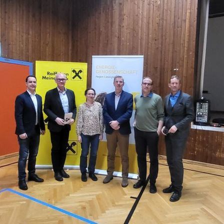 Sechs Personen stehen nebeneinander und posieren für ein Foto. Sie sind alle geschäftlich gekleidet. Hinter ihnen befindet sich ein gelber Banner mit schwarzen Buchstaben.