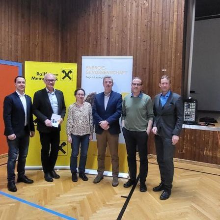 Fünf Personen stehen in einem Raum, alle in formeller Kleidung. Sie posieren für ein Foto vor einer Holzwand mit gelben Bannern. Ein Mann hält ein Stück Papier.