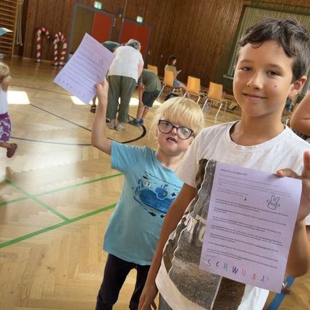 Eine Gruppe von Kindern mit Papieren steht in einer Turnhalle. Ein Junge mit Brille und ein Junge mit einem hellblauen T-Shirt posieren für ein Foto.