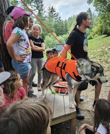 Eine Gruppe von Kindern und Erwachsenen versammelt sich um einen Hund in einem orangefarbenen Schwimmweste, der auf einer Holzplattform steht, in einer Outdoor-Umgebung.