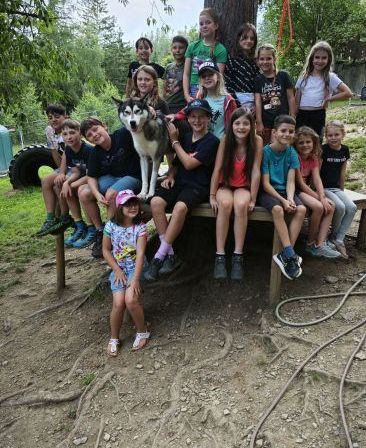 Eine Gruppe von Kindern und einem Hund sitzen auf einer Holzbank in einem Park. Einige von ihnen tragen Hüte und Schuhe. Der Hund steht vor einem Baum mit blauen Bändern.