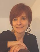 Eine Frau mit kurzen roten Haaren lächelt und posiert für ein Foto. Sie trägt ein schwarzes Oberteil und hat Ohrringe mit Spiraldesign.