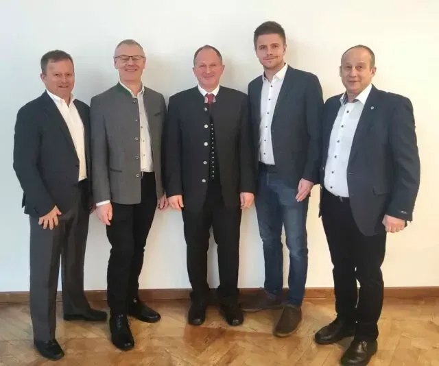 Fünf Männer, gekleidet in formeller Kleidung, stehen in einer Reihe vor einer schlichten weißen Wand und scheinen für ein Foto zu posieren.