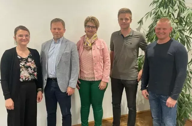 Fünf Menschen stehen vor einer weißen Wand. Die Frau links trägt eine Brille und ein Blumenoberteil. Die Frau in der Mitte trägt einen Schal. Der Mann rechts trägt ein dunkelblaues Shirt.