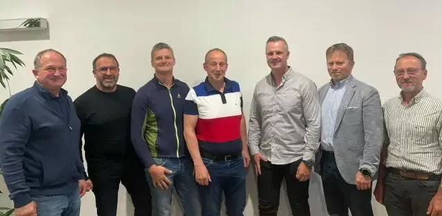 Sieben Männer stehen nebeneinander vor einer weißen Wand und lächeln für ein Foto.