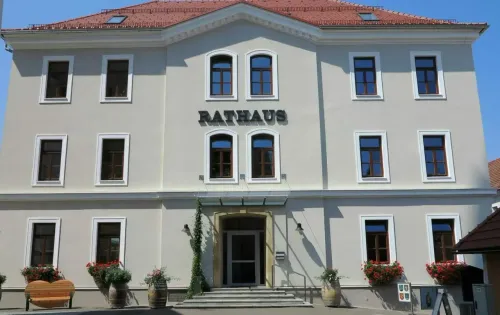 Ein weißes Gebäude mit einem roten Dach, namens Rathaus, hat mehrere Fenster und einen zentralen Eingang. Der Eingang wird von Treppen und Topfpflanzen flankiert.