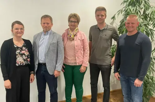 Fünf Personen posieren für ein Foto und stehen eng beieinander in einem Raum. Sie lächeln. Die Frau trägt einen Schal und der Mann rechts trägt eine Uhr. Hinter ihnen steht eine Pflanze.