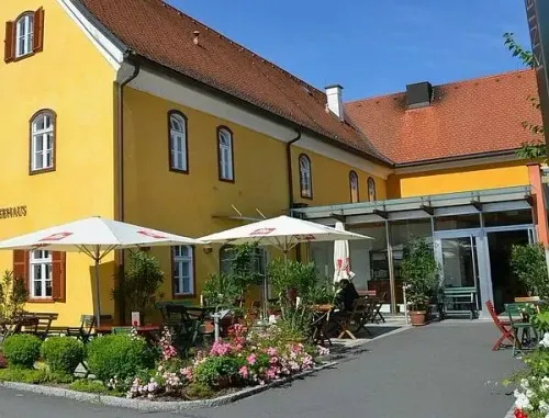 Ein gelbes Gebäude mit braunem Dach hat mehrere Fenster und Pflanzen draußen. Es gibt weiße Sonnenschirme, Tische und Stühle. Eine Person sitzt auf einem Stuhl unter dem Schirm.