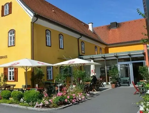 Ein gelbes Gebäude mit braunem Dach hat mehrere Fenster und Pflanzen draußen. Es gibt weiße Sonnenschirme, Tische und Stühle. Eine Person sitzt auf einem Stuhl unter dem Schirm.