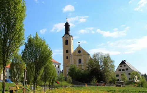 Eine gelbe Kirche mit einem Glockenturm steht auf einem Feld mit Bäumen und einem Gebäude dahinter. Der Himmel ist klar mit Wolken.