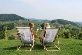 Zwei Frauen sitzen auf Holzstühlen in einem Weinberg, halten Weingläser in den Händen und genießen die Aussicht auf ein grünes Tal und entfernte Häuser.