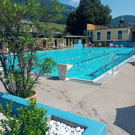 Ein Freibad umgeben von Bäumen und Pflanzen, mit einem Gebäude und Bergen im Hintergrund.
