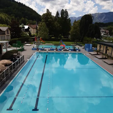 Ein Freibad mit Bahnen, umgeben von Bäumen und Bergen, mit einem Gebäude und einer Rutsche in der Ferne.