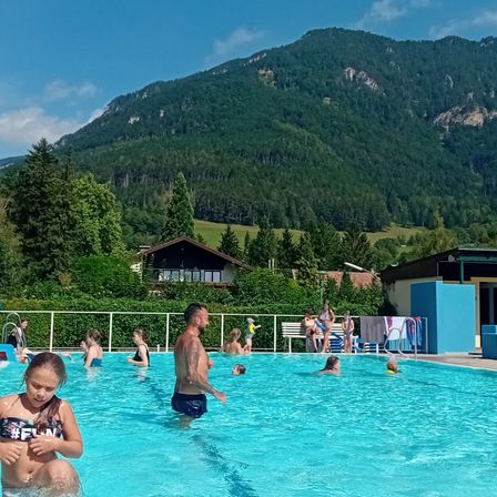 Freibad mit einer Gruppe von Menschen, die sich an einem sonnigen Tag amüsieren, mit Bergen im Hintergrund.