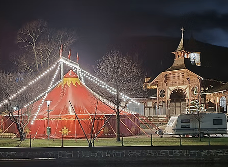 Ein Zirkuszelt ist auf einem Rasenfeld aufgebaut und bei Nacht von Lichtern beleuchtet. In der Nähe befinden sich ein Wohnwagen und ein Gebäude mit Glockenturm.