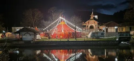 Ein leuchtendes Zirkuszelt wird von Lichtern erhellt und am Fluss positioniert, mit einem Pavillon, Gebäuden und Bergen im Hintergrund.