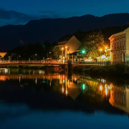 Eine Nachtansicht einer Stadt mit Gebäuden, einer Brücke und einem Fluss, der Lichter reflektiert. Im Hintergrund sind Berge zu sehen, und der Himmel ist dunkel.