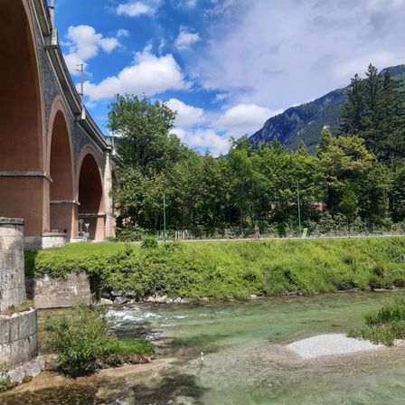 Ein Fluss fließt unter einer historischen Brücke mit Bogenöffnungen, umgeben von üppigem Grün und Bergen in der Ferne an einem sonnigen Tag.