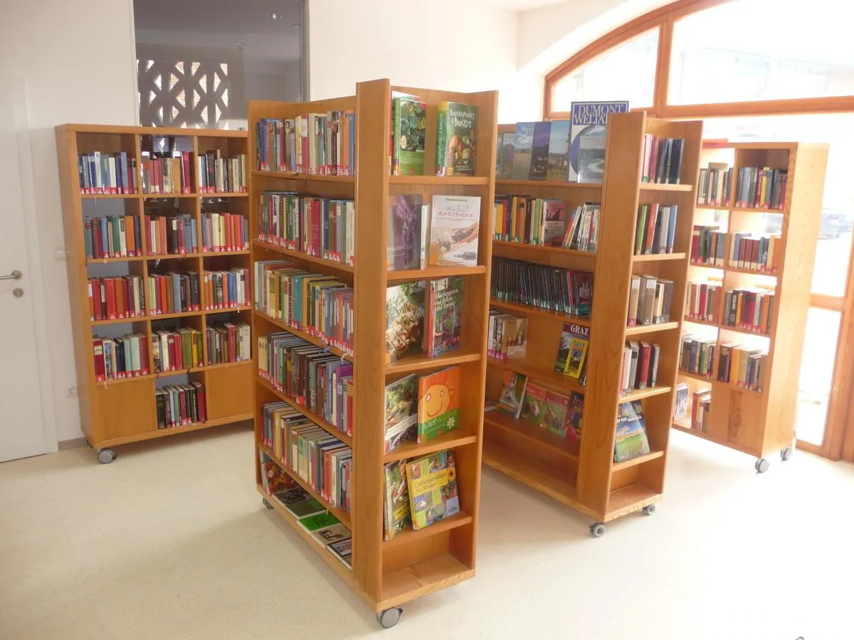 Eine Innenbibliothek mit Reihen von Holzregalen, die mit verschiedenen Büchern gefüllt sind. Die Bücherregale sind in einer Reihe mit einem Abstand zwischen ihnen angeordnet. Der Boden ist weiß und der Raum hat Glasfenster.