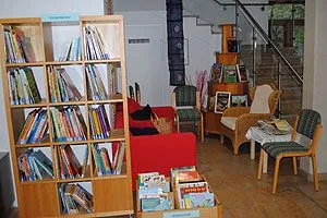 Ein Wohnzimmer mit einem Bücherregal voller Bücher, einem roten Sofa, einem Korbsessel und einem kleinen Tisch mit einem Korb darauf. Im Hintergrund befindet sich eine Treppe mit einer Metallbrüstung.