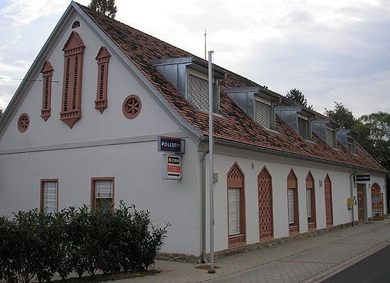 Ein weißes Gebäude mit einem roten Dach, Ziegeldetails und einem Schild, das 'POLIZEI' steht. Das Gebäude hat gewölbte Fenster und Türen mit Holzläden. Es befindet sich an einer Straße mit Gehweg und Büschen.