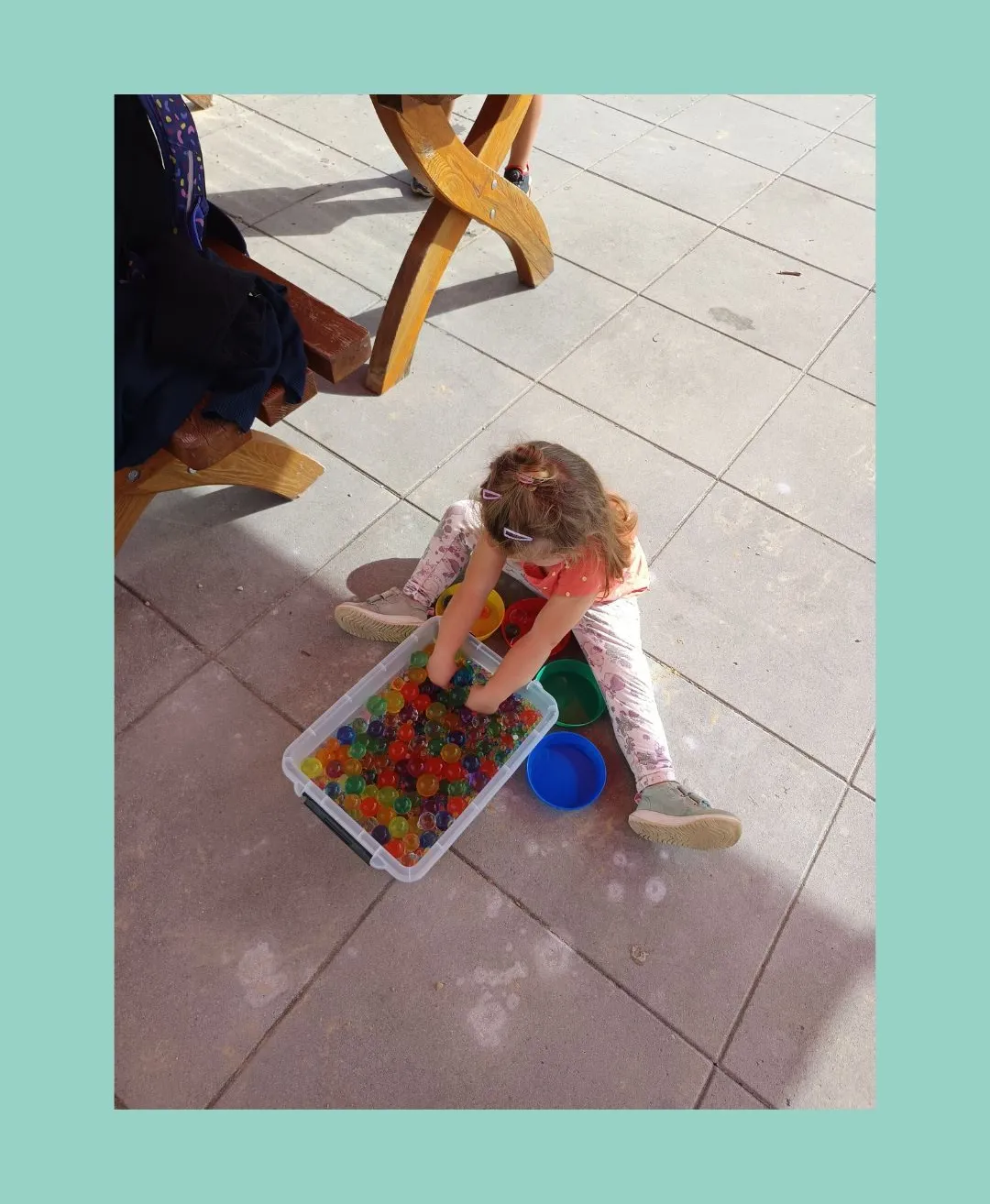 Ein kleines Mädchen spielt mit bunten Murmeln in einem Plastikbehälter auf einem gefliesten Boden.