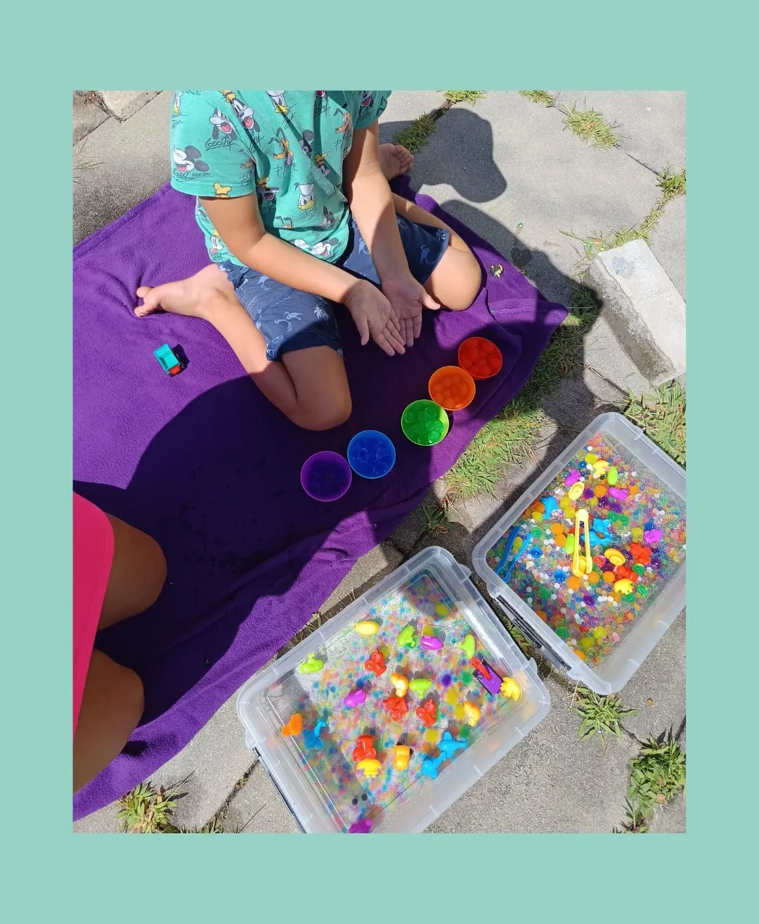 Ein Kind sitzt auf einem lila Handtuch mit einem Spielzeugauto und Plastikbehältern voller bunter Gegenstände.