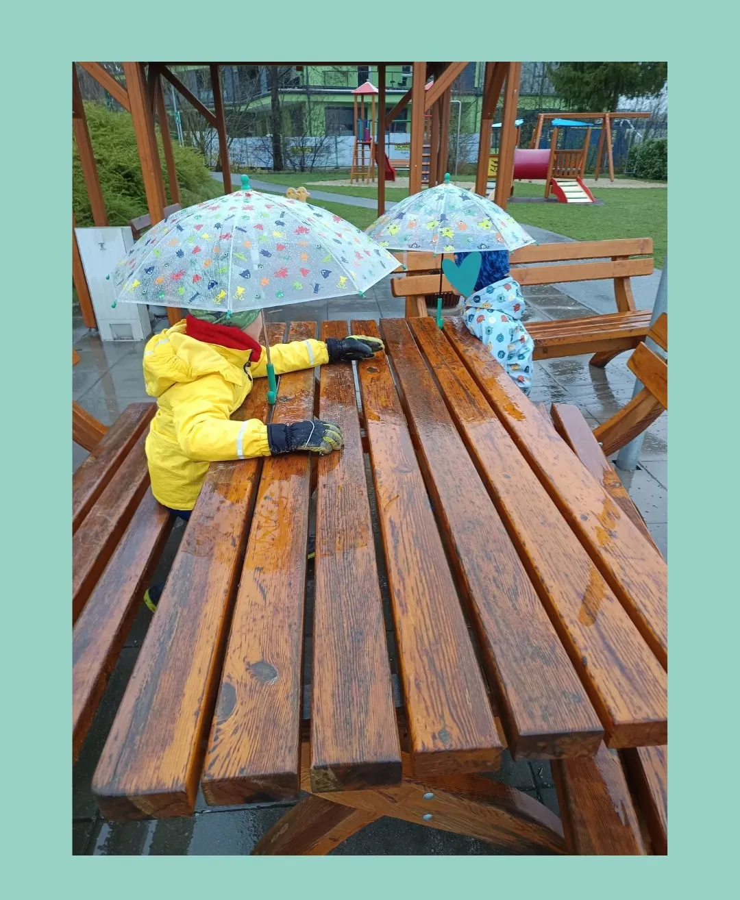 Zwei Kinder sitzen an einem hölzernen Picknicktisch in einem Park und halten bunte Regenschirme. Sie tragen gelbe Jacken und Handschuhe, mit einer Bank und einem Spielplatz im Hintergrund.