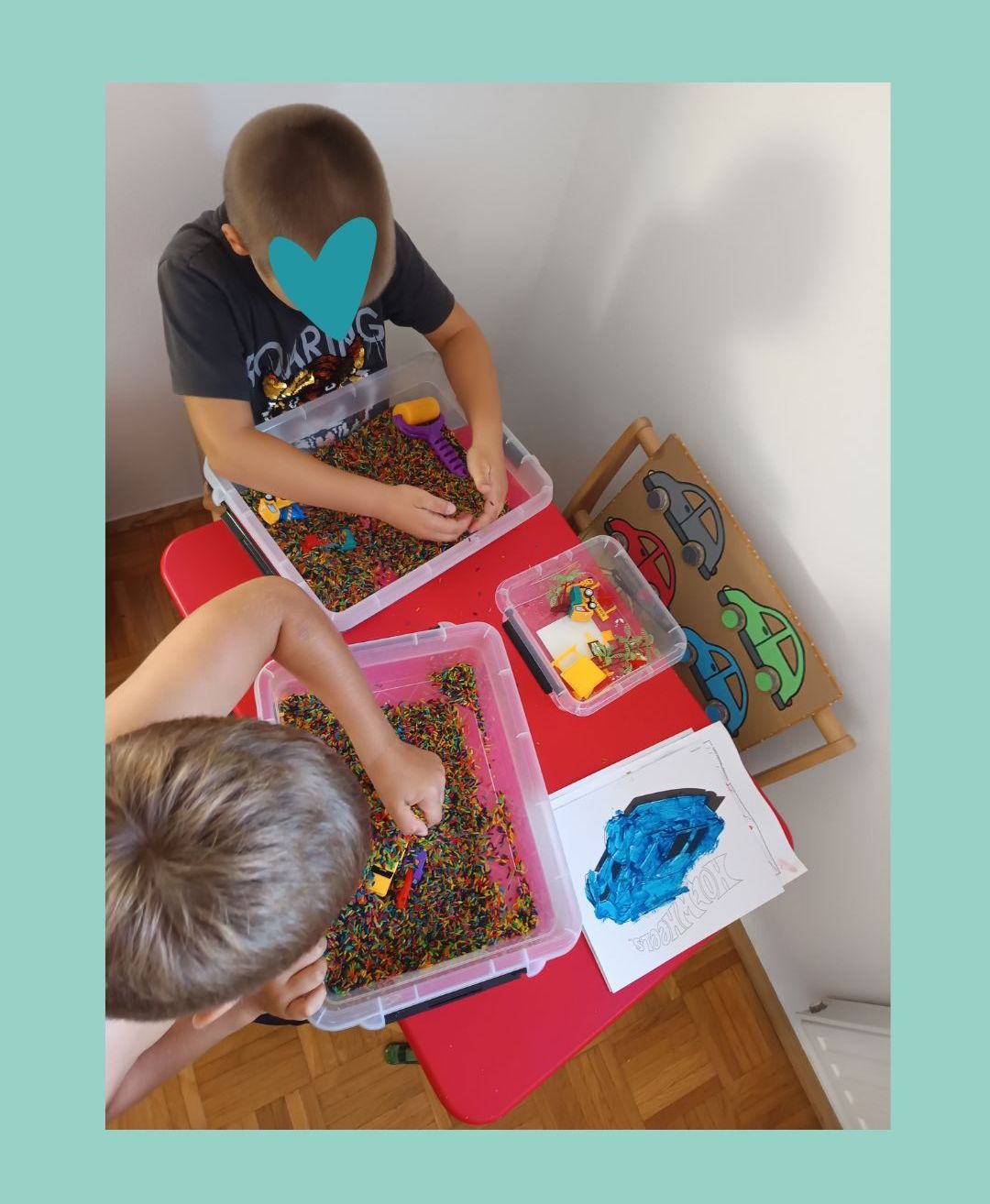 Zwei junge Jungen spielen mit Spielzeug in durchsichtigen Plastikbehältern auf einem roten Tisch. Ein Kind hat ein blaues Herz auf dem Gesicht. Es gibt andere Spielzeuge, einschließlich Autos, die auf dem Tisch verstreut sind.