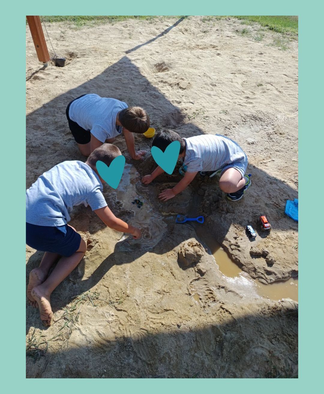 Drei Jungen spielen im Sand und graben jeweils mit ihren Händen und Spielzeug. Sie befinden sich in einem schattigen Bereich, mit Spielzeug und einem gelben Eimer in der Nähe.