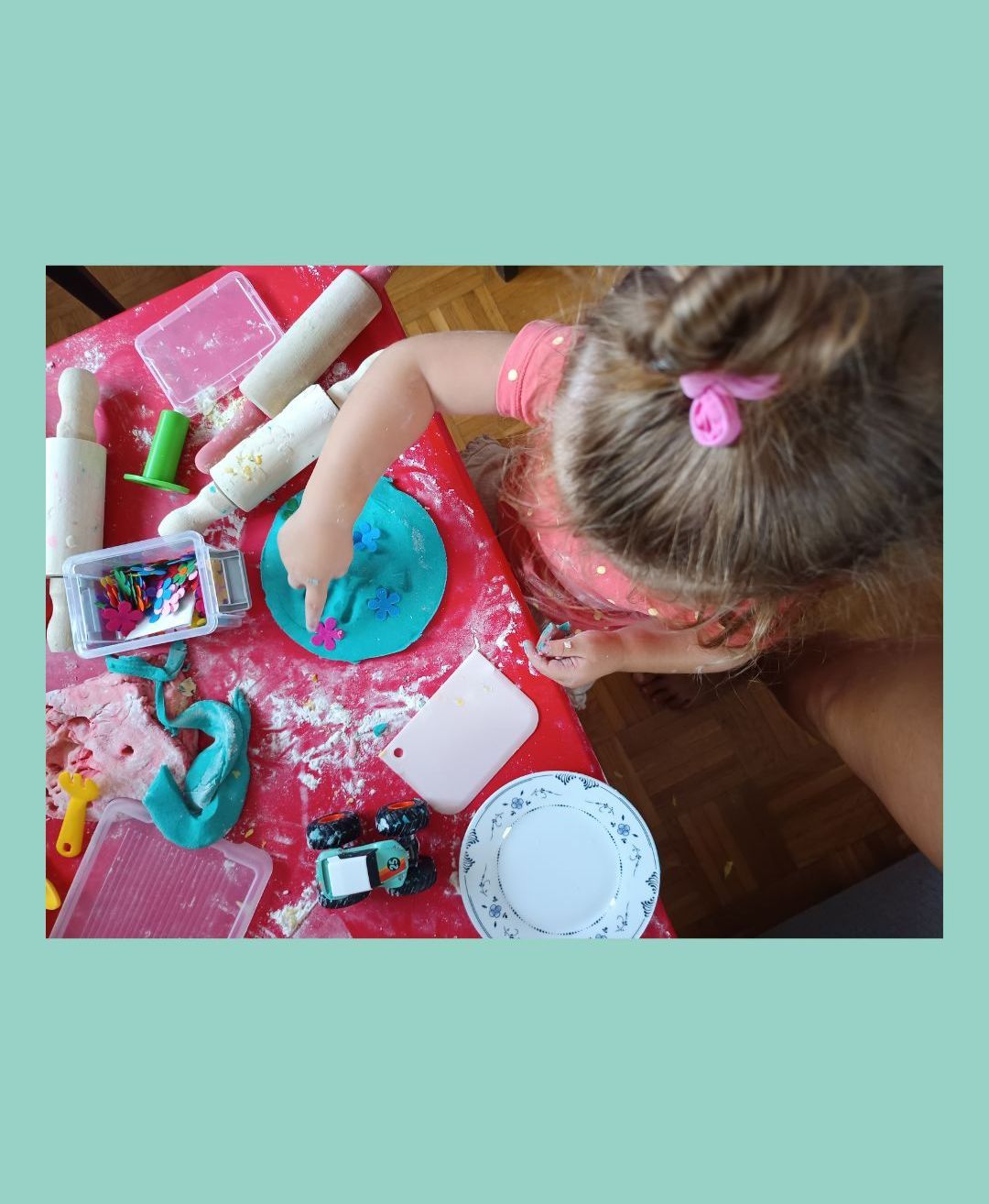 Ein Kind spielt mit Modelliermasse auf einem roten Tisch, umgeben von verschiedenen Werkzeugen und Spielzeug, einschließlich eines Tellers und Nudelholze.