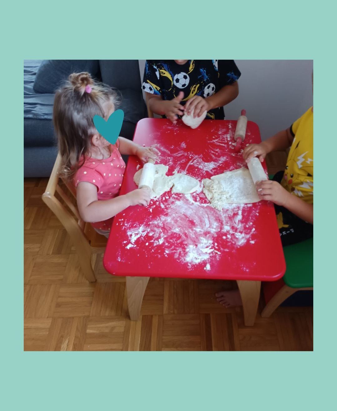 Drei Kinder spielen mit Teig auf einem roten Tisch. Das Mädchen in Pink hat einen Herzaufkleber auf der Wange.