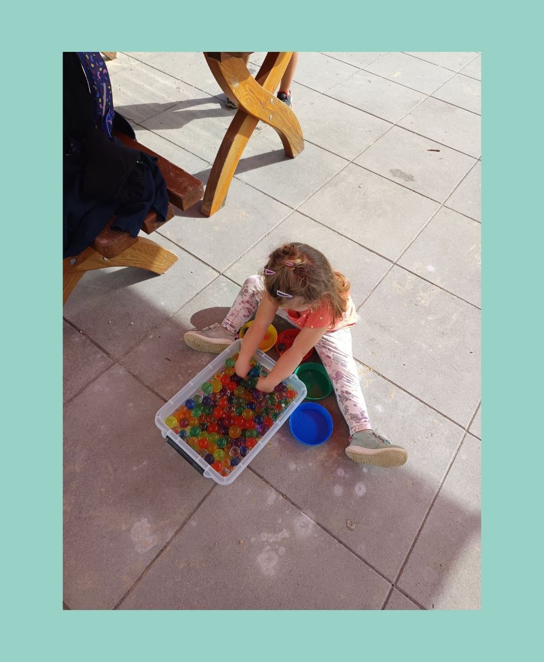 Ein kleines Mädchen spielt mit bunten Murmeln in einem Plastikbehälter auf einem gefliesten Boden.