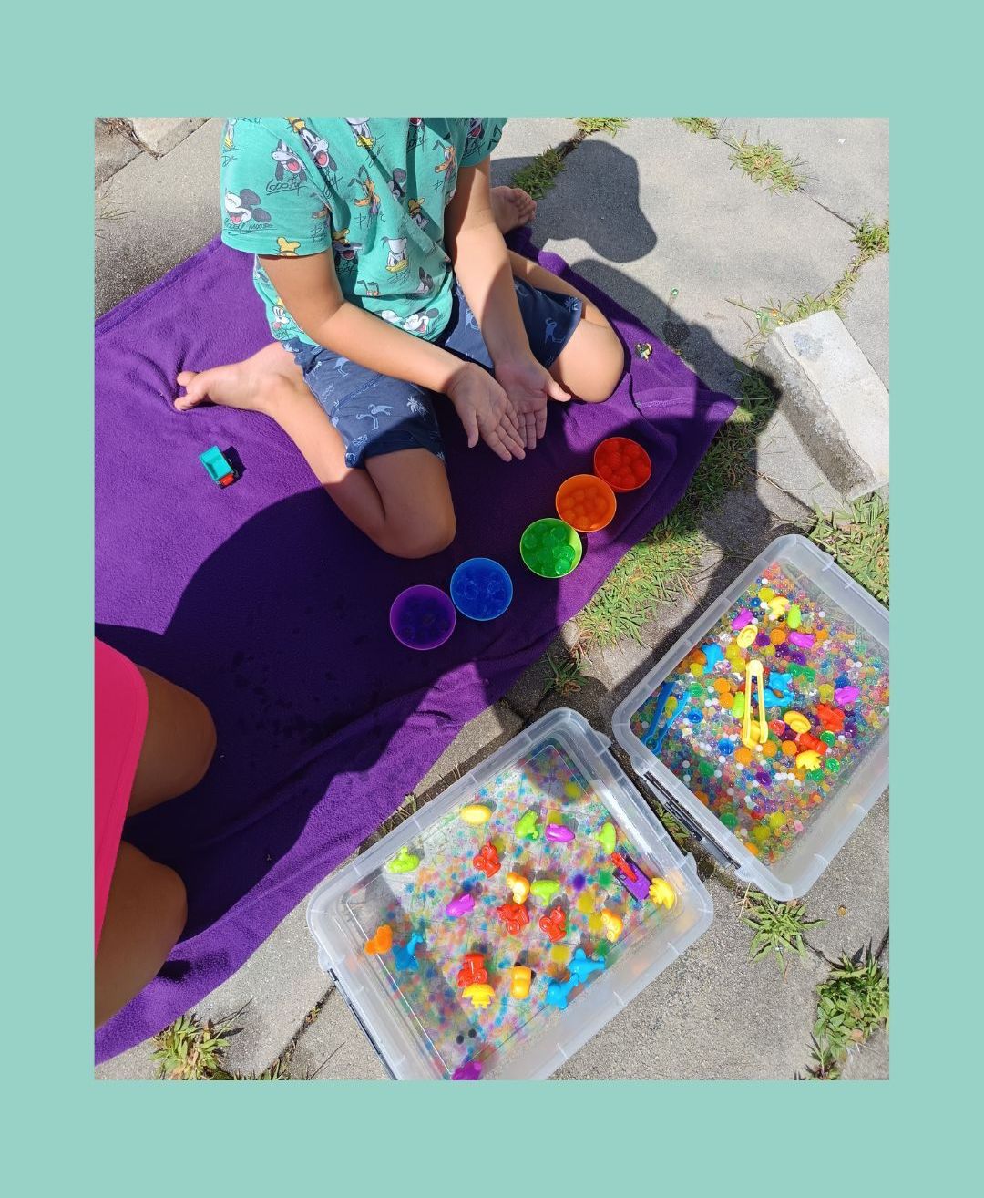 Ein Kind sitzt auf einem lila Handtuch mit einem Spielzeugauto und Plastikbehältern voller bunter Gegenstände.