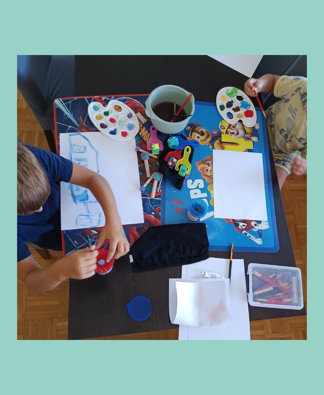 Zwei Kinder sitzen an einem Tisch mit einem Kunstprojekt, jedes hält einen Pinsel. Verschiedene Malutensilien, einschließlich Farbe, Pinsel und eine Tasse, sind auf dem Tisch. Ein Kind zeichnet auf Papier, während das andere ein rotes Objekt hält.