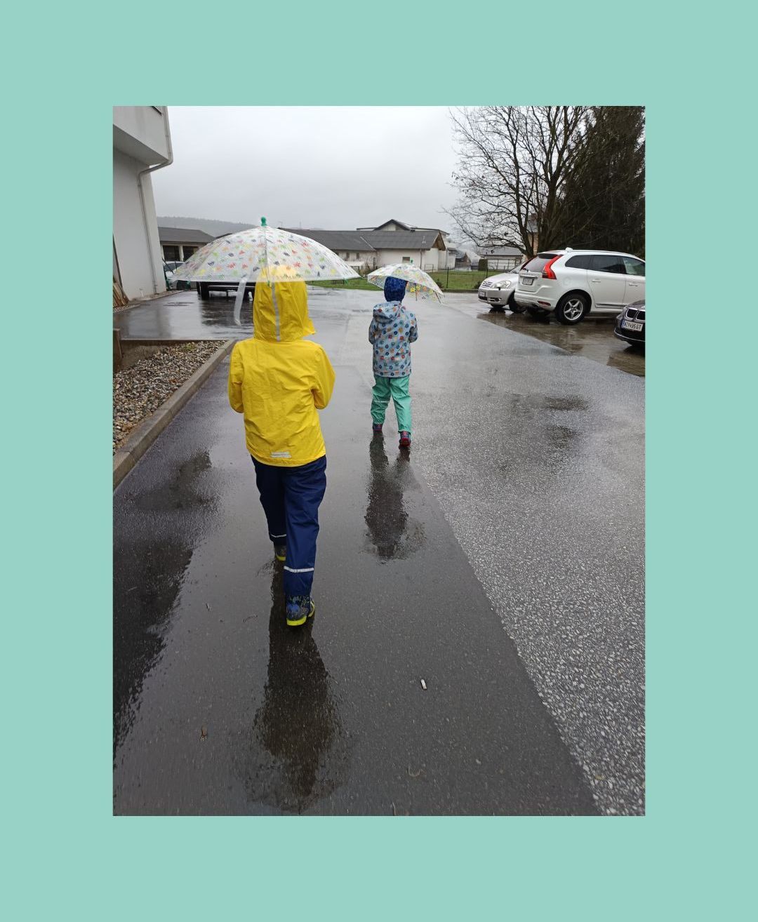 Zwei Kinder in Regenmänteln und Stiefeln gehen auf einer nassen Straße. Eines hält einen Regenschirm, das andere einen bunten. Autos sind am Straßenrand geparkt.