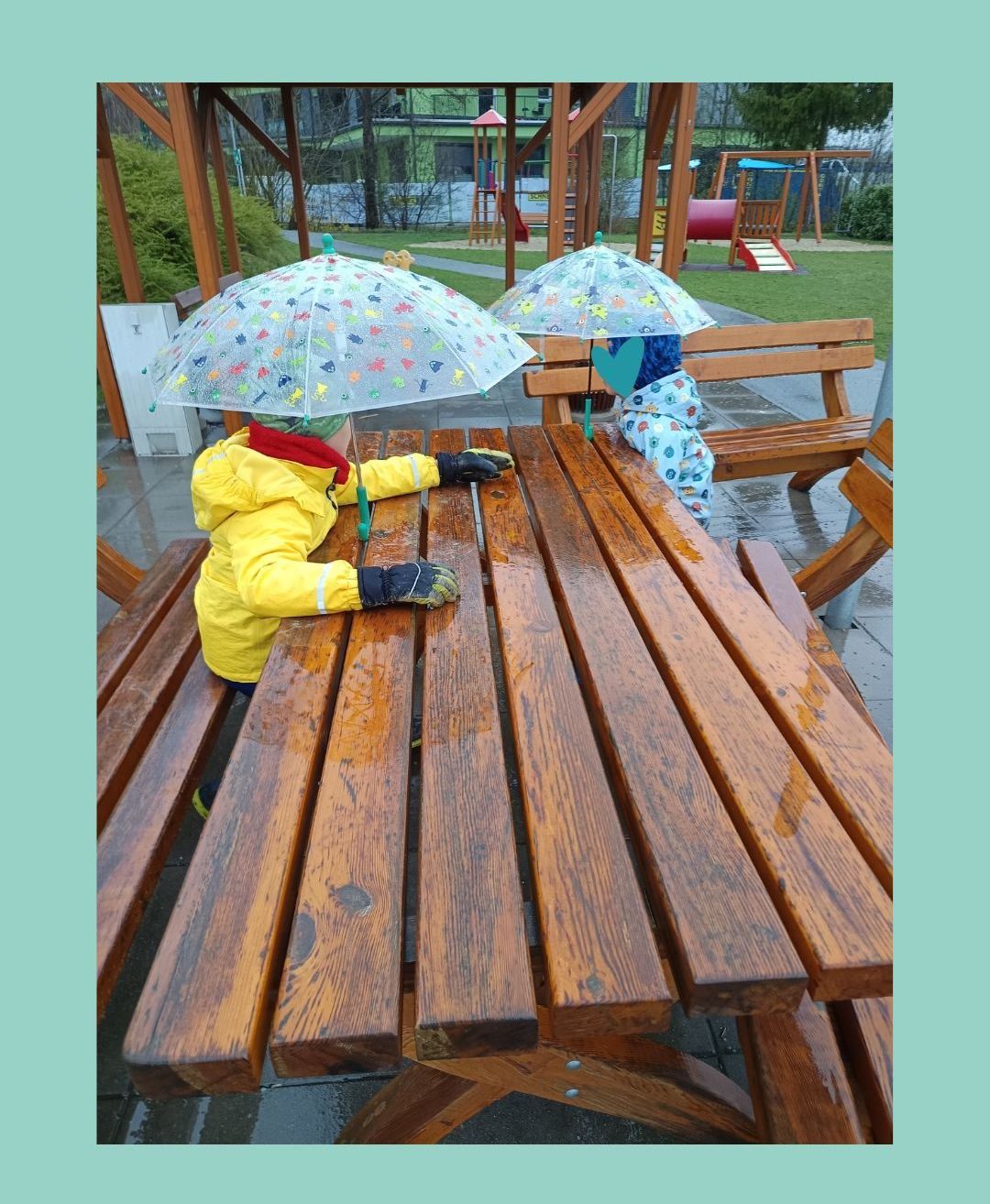 Zwei Kinder sitzen an einem hölzernen Picknicktisch in einem Park und halten bunte Regenschirme. Sie tragen gelbe Jacken und Handschuhe, mit einer Bank und einem Spielplatz im Hintergrund.